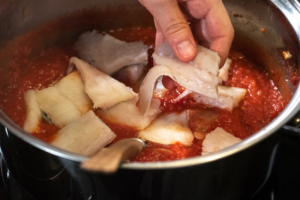 Fish pieces being put into tomato-based stew after being cut into chunks